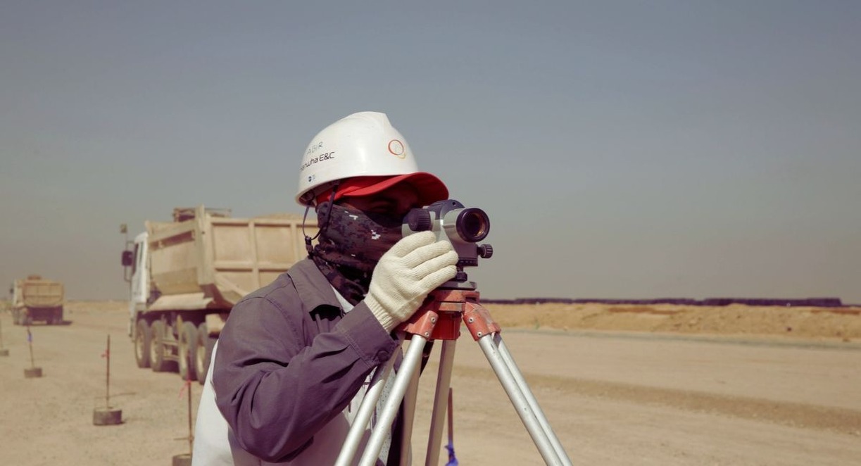 Géomètre en casque de chantier visant avec un niveau optique sur trépied, camions-bennes en arrière-plan